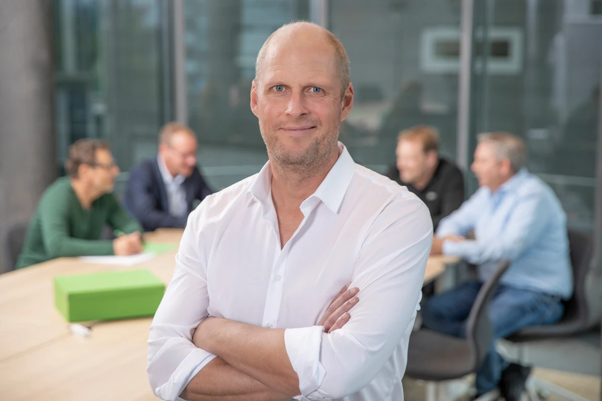 Joschi Alger steht mit verschränkten Armen in einem modernen Konferenzraum und blickt selbstbewusst in die Kamera. Im Hintergrund sitzt eine Gruppe im Gespräch.